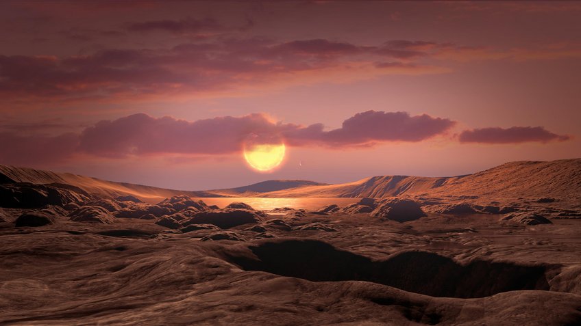 Artist’s conception of a rocky Earth-mass exoplanet like Wolf 1069 b orbiting a red dwarf star. If the planet had retained its atmosphere, chances are high that it would feature liquid water and habitable conditions over a wide area of its dayside. Surface of a fictitious planet. It consists of hills of rock. In the background is a lake. The sky has some clouds. Near the horizon a sun shines and bathes the picture in a reddish colour.