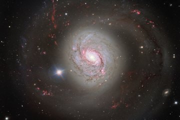 A vivid image of the spiral galaxy Messier 77 (M77), also known as NGC 1068. The galaxy exhibits an impressive structure with a brightly glowing centre and spiralling arms extending far into space. The spiral arms are highlighted by pink and red regions, representing areas of active star formation. The galaxy is surrounded by countless small points of light, representing distant stars and galaxies. In the foreground, a bright, star-like point shines on the left side of the galaxy. The galaxy's colours range from white and blue in the centre to reddish hues in the outer regions.
