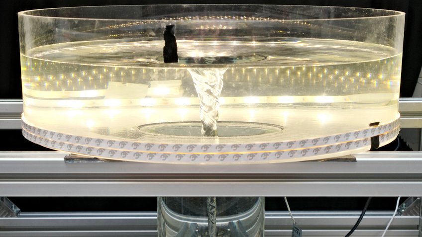 Dieses Foto zeigt den oberen Bereich des Wasser-Tornado-Modells. Das Becken aus Plexiglas hat einen Durchmesser von 50 Zentimetern und wird mit LED-Streifen beleuchtet. Das Wasser darin entwickelt einen Wirbel, dessen Oberfläche das Profil eines Gravitationsfelds nachbildet. Die Auswertung hat ergeben, dass sich die Bewegungen des Wassers ähnlich wie diejenigen in einer protoplanetaren Scheibe verhalten. Ein zylindrisches, transparentes Gefäß steht auf einer offenen, silberfarbenen Metallkonstruktion. Es ist mit klarem Wasser gefüllt, in dessen Zentrum ein schlanker, trichterförmiger Wirbel von der Oberfläche bis zum Boden rotiert. Die Wasseroberfläche ist in der Mitte eingezogen und an den Rändern leicht wellig. Kleine Luftbläschen werden vom Wirbel nach unten transportiert und ziehen feine Linien entlang der Gefäßwand. Rund um das Gefäß verlaufen LED-Lichtbänder, die den Wirbel kontrastreich aus der dunklen Umgebung hervorheben. Die Szene wirkt wie eine wissenschaftliche Demonstration, bei der die Dynamik von Wasserströmungen anschaulich gemacht wird.