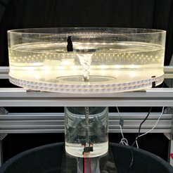 Fotografie eines Wasser-Tornado-Modells in einem abgedunkelten Labor. Im Zentrum steht eine silberfarbene Metallkonstruktion, auf der ein kreisrundes, transparentes Plexiglas-Becken mit 50 cm Durchmesser ruht. Die Außenwand des Beckens ist mit schmalen weißen LED-Streifen versehen, die von unten nach oben gleichmäßig leuchten. Unter dem breiten Becken schließt sich ein engerer Zylinder an, dessen glatte Wände nach unten bis zu zwei schwarzen Düsen führen. An diesen Düsen sind Schläuche angeschlossen, die kontinuierlich Wasser einspeisen. Durch den Wasserzufluss bildet sich ein schlanker, trichterförmiger Wirbel, der von der Oberfläche bis zum Boden reicht und im Profil an ein Gravitationsfeld erinnert. Mehrere Kabel und Schläuche verlaufen entlang der Metallkonstruktion. Der helle Wasserwirbel hebt sich kontrastreich vom dunklen Hintergrund ab und vermittelt den Eindruck eines präzise kontrollierten wissenschaftlichen Experiments.