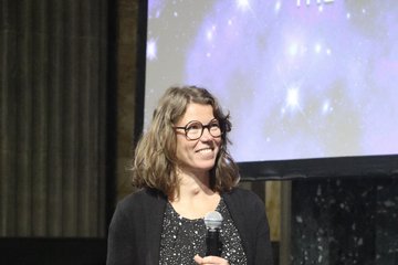 Eine Frau, nämlich Ruth Grützbauch, steht lächelnd auf einer Bühne. Sie hält eine Medaille und ein Stück Papier in der Hand.