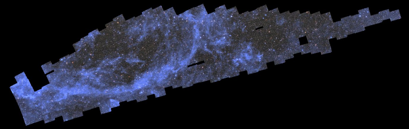 A wide, elongated black background with a long blueish patched-up field of stars, galaxies, and wispy clouds in the upper left diagonal of the image. This patchy mosaic consists of multiple almost square photographs arranged in a shape that loosely resembles a thin, pixelated whale diving towards the bottom left of the image. The myriad of sparkling galaxies and stars in this mosaic crowd together to create the impression of a dark grey field with frequent speckles of bright pastel yellow, red and blue dots of light at an even distribution. A streak of silver-blue diffuse clouds in a half-elliptical shape in the left half of the image and a small round patch just right of the centre stand out particularly. There are small rectangular holes in the mosaic at three spots right of the image centre. The pitch-black background is peaking through and starkly contrasts the sparkling image field.