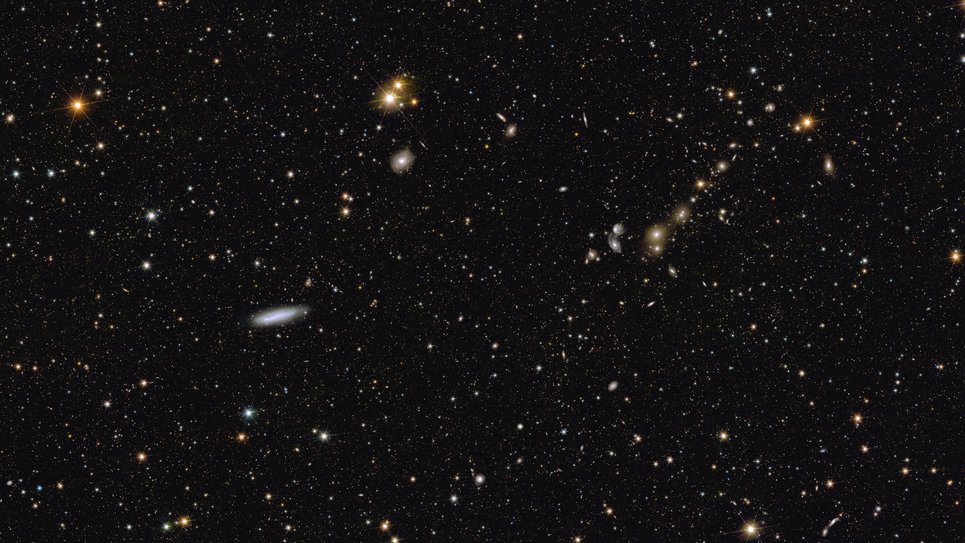 A sea of light points scattered evenly across a black background, with a few brighter and colourful ones standing out. As one gazes across the image from left to right, a bright orange star with six faint spikes catches the eye first in the upper left corner. To the lower right of this, just below the picture's halfway mark, a thin white elliptical galaxy floats into sight. Above it slightly to the right, a pair of bright golden yellow stars dance into view. Further to the right, in the last third of the image, a short line of stars and galaxies is visible. This appears as a chain, tilted at a 45-degree angle, on which hazy galaxies in different hues of yellow are strung like beads on a cosmic wristband.