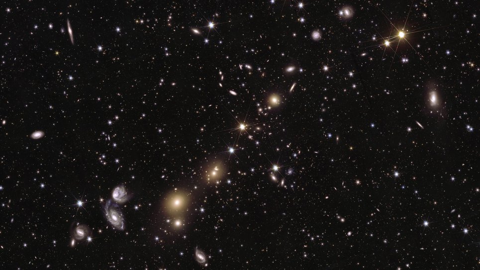 A dark black backdrop with bright dots of stars and disks of galaxies scattered across it. Fading into the background are myriad light points, like specks of white paint and light hues of yellow, blue, and purple. In the foreground, a handful of bright light sources draw attention. Spanning from the top right corner to the bottom left of the image, yellow light points form a diagonal string of sparkling beads. Some of these are sharp, piercing dots with six faint spikes; others are blurry blobs of light, gleaming bright in their centre with their edges blending into the depths of the black background. In the lower left quarter of the image, two hazy white spiral shapes of light catch the eye, which appear to be swirling around each other.
