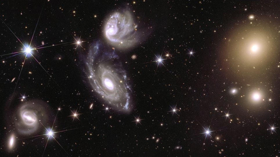 Three groups of light sources and a scatter of piercing dots of light with six faint spikes stand out in stark contrast against a black backdrop. The most prominent light sources occupy the centre of the image. They are two hazy white spirals, that appear to be swirling in a cosmic dance with each other, with the lower spiral being larger than the one above it. On the right side of the image, two spots of gleaming yellow light draw attention. The hazy light blobs emit a golden glow from their centre, which fades out in a circular shape into the background. Another spiral shape can be seen in the bottom left corner of the picture. It appears as if it is a thin white bar spinning in a circle and emitting a white spray of paint at its ends, leaving behind a diffuse trace of light.