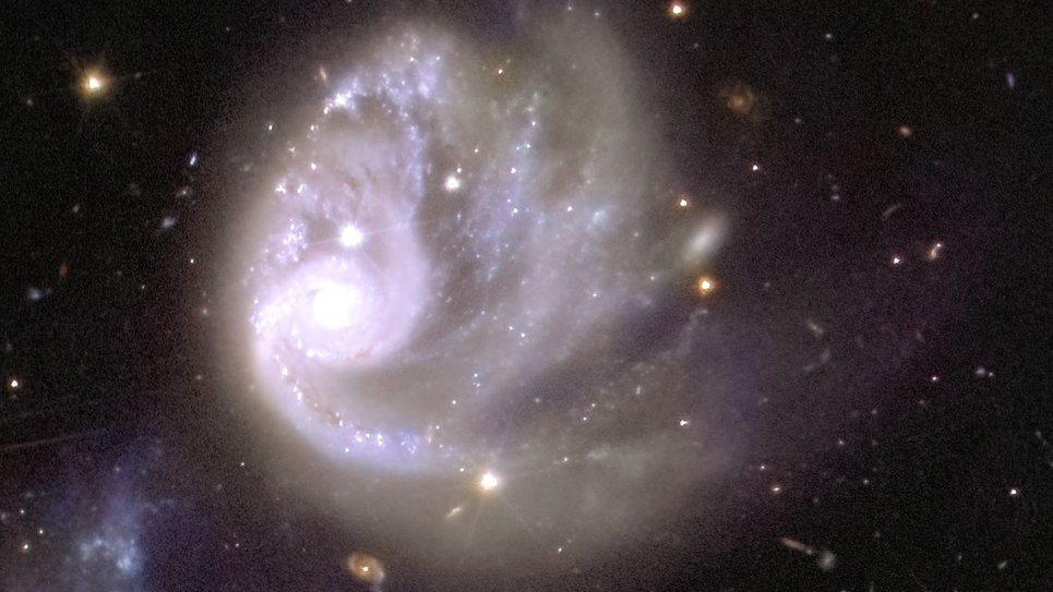 The centre of the image shows a large white swirl of light, framed by a sea of blackness and a few individual dots and smears of colour in the background. Off-centre to the left of this spiral sits a gleaming circle, the brightest spot of the image. Starting at this white ball of light, sparkling arches trace the shape of a tornado rotating clockwise, spewing glitter into empty space across the image.