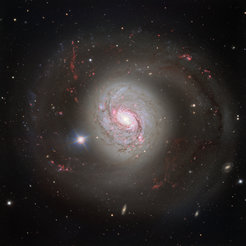 A vivid image of the spiral galaxy Messier 77 (M77), also known as NGC 1068. The galaxy exhibits an impressive structure with a brightly glowing centre and spiralling arms extending far into space. The spiral arms are highlighted by pink and red regions, representing areas of active star formation. The galaxy is surrounded by countless small points of light, representing distant stars and galaxies. In the foreground, a bright, star-like point shines on the left side of the galaxy. The galaxy's colours range from white and blue in the centre to reddish hues in the outer regions.