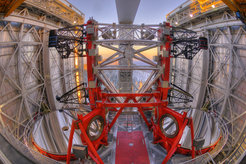 The image shows the interior of the Large Binocular Telescope (LBT), one of the world's most powerful telescopes. At the centre are two large, round, highly reflective primary mirrors arranged in parallel. The mirrors are surrounded by a massive red steel structure with a geometrically intricate design, supported by diagonal and rectangular braces for stability. Above the mirrors, two black arm-like structures with cylindrical components extend outward, likely housing instruments or sensors. The telescope’s roof is open, revealing the surrounding symmetrical metal framework with its curved and rectangular elements. In the background, a snow-covered forest landscape stretches under a sky glowing with soft orange and blue hues. The scene blends technical precision with aesthetic harmony, offering a captivating glimpse into the geometric elegance of this research instrument.