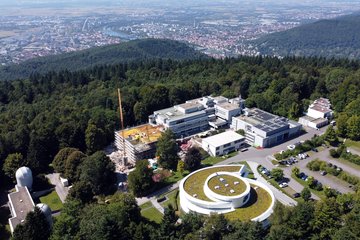 Aerial view of the campus of the Max Planck Institute for Astronomy (MPIA) on the Königstuhl hill in Heidelberg. In the foreground at the lower right stands the Haus der Astronomie, a modern white building in a spiral shape with a green roof. To its left are several additional institute buildings with flat roofs, some fitted with solar panels, and a new construction site with a crane and yellow scaffolding. In the lower left, two white domes of small observatories are visible. Parking areas with cars are located in front of the buildings. The entire campus is surrounded by dense green forest. In the background stretches the Neckar valley with the city of Heidelberg, the river with bridges, and the wide Upper Rhine Plain extending to the horizon.